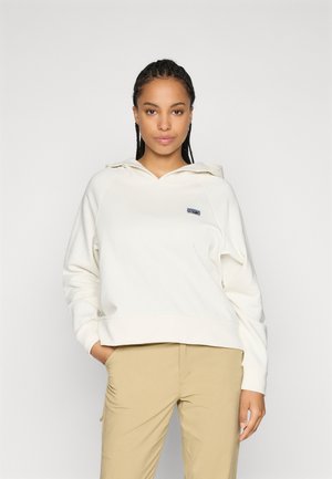 Jonge vrouw met gevlochten haar, die een witte sweatshirt met capuchon en beige broek draagt. Ze staat met een hand in haar zak tegen een effen achtergrond.