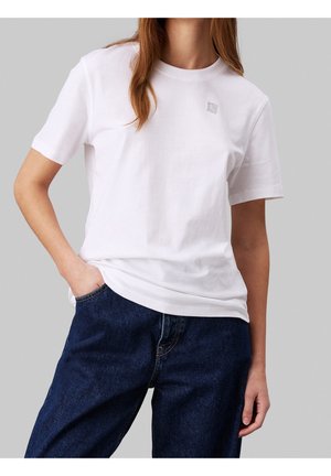 Osoba ubrana w gładki biały t-shirt z krótkim rękawem z małym logo na piersi i ciemnoniebieskie dżinsy, stojąca z jedną ręką w kieszeni.
