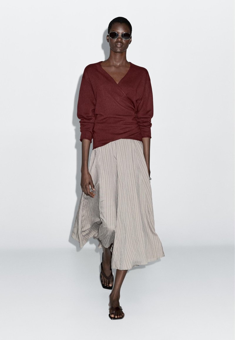 Bordeauxrode trui met een wikkelontwerp, gecombineerd met een licht gestreepte midi-rok. Model draagt donkere zonnebril en platte sandalen. Gladde stoffen.