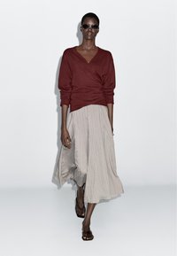 Vinrød sweater med omslagsdesign, kombineret med et lyst, stripet midiskørt. Modellen bærer mørke solbriller og flade sandaler. Glatte stoffer.