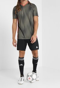 Svarta fotbollsbyxor, mörkgrön strukturerad sporttröja med röda detaljer, svarta strumpor med vita ränder och en vit fotboll med svarta mönster.