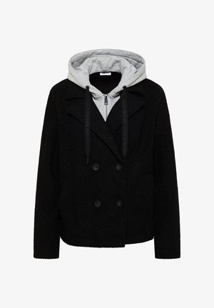 Cappotto nero doppiopetto in tessuto sovrapposto a una felpa grigia con zip e cappuccio, dotata di lacci neri.
