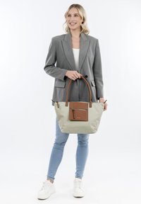Beige tote bag with brown leather accents, vertical zipper pocket, and sturdy round handles. Model wears a gray blazer and jeans.