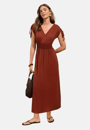Kvinde iført rustfarvet V-hals kjole med rynket talje, bærende en flettet håndtaske og iført sandaler og markante øreringe.
