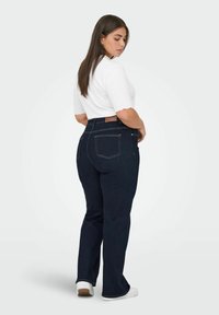 Augsta jostas līmeņa tumši zili džinsi ar taisnu kāju, redzamām aizmugurējām kabatām un gaišu šuvju. Apvienoti ar baltu ribotu T-kreklu ar īsām piedurknēm.