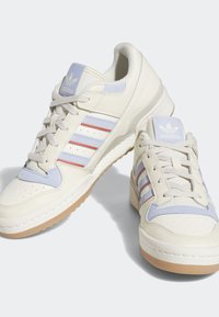 Zapatillas Adidas con un upper de cuero blanco crema, acentos en azul, tres rayas rojas y azules, y suela de goma.
