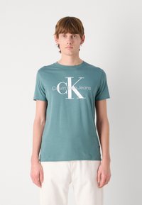 Žaliojo medvilnės marškinėliai su „Calvin Klein Jeans“ tekstu ir logotipu balta spalva. Trumpi rankovės, apvalus kaklas ir reguliari forma. Minimalistinis dizainas.