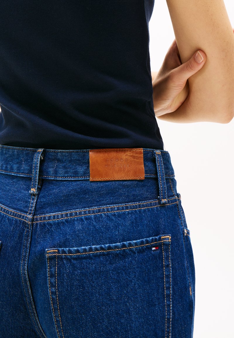 Niebieskie dżinsy z denimu o strukturalnym wykończeniu, z brązową skórzaną łatą z napisem "TOMMY HILFIGER" na tylnym pasku w talii i kontrastowymi przeszyciami.