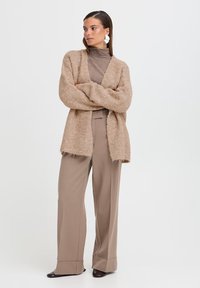 Cardigan beige morbido con apertura frontale, maniche lunghe e bordi sfrangiati, abbinato a pantaloni marroni a gamba larga e tacchi scuri.
