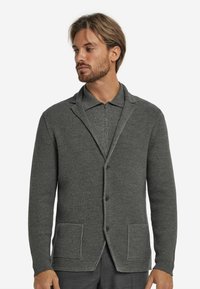 Grau strukturierte Strickjacke mit einem Schalkragen, zwei Vordertaschen und Knopfverschlüssen. Gerippter Stoff. Über einem grauen Reißverschlusshemd getragen.