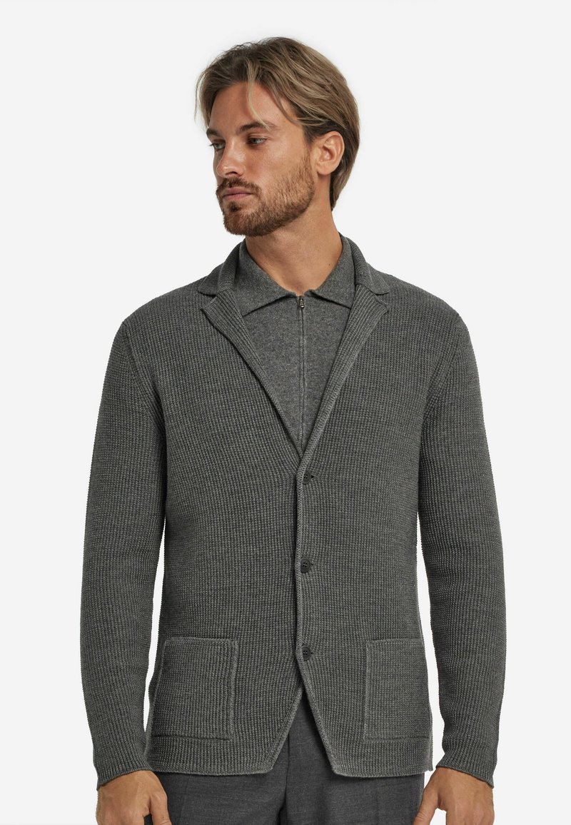 Grau strukturierte Strickjacke mit einem Schalkragen, zwei Vordertaschen und Knopfverschlüssen. Gerippter Stoff. Über einem grauen Reißverschlusshemd getragen.