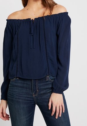 Mujer con una blusa azul marino de manga larga y hombros descubiertos, combinada con jeans de mezclilla azul oscuro, de pie con una mano visible.