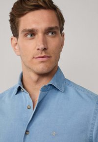 Joven con cabello castaño claro que lleva una camisa de botones azul claro con un pequeño logo bordado en el pecho, mirando hacia un lado.