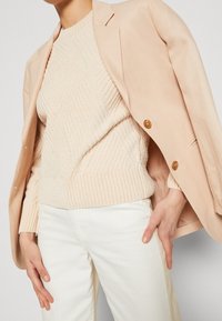 Person som bär en strukturerad beige tröja, en ljusbeige kavaj över axlarna och vita byxor, med ena handen vilande på höften.