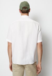 Chemise blanche à manches courtes en lin avec un ourlet arrondi, associée à un pantalon beige clair. Une casquette verte en coton apporte du contraste.