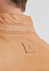 Der Kragen einer cognacfarbenen Lederjacke zeigt eine strukturierte Oberfläche, sichtbare Nähte und ein rechteckiges Patch mit einem eingeprägten Sterndetail.