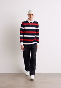 Stribet langærmet polo i navy, rød og hvid, parret med mørke jeans og hvide sneakers. Modellen bærer en lysfarvet kasket.