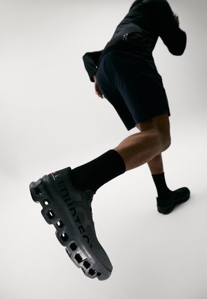 Čierne športové topánky s sieťovinovým zvrškom, hrubou podrážkou s viditeľnými výrezmi a logom na boku. Nosené s čiernymi ponožkami a šortkami.