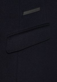 Granatowy płaszcz z wełny z elegancką, płaską kieszenią z czarnym wykończeniem oraz subtelnym brązowym przeszyciem.