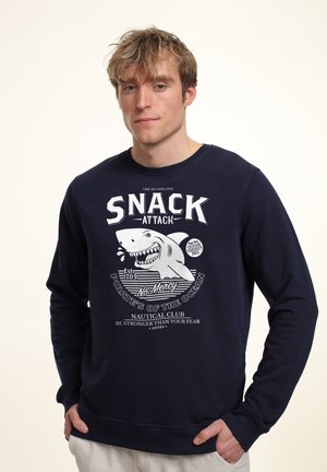 Navy blue sweatshirt made of cotton, featuring a shark graphic and text "SNACK ATTACK," with ribbed cuffs and a relaxed fit.