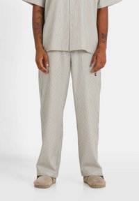 Ensemble chemise et pantalon en tons beige et crème. Tissu léger, manches courtes, coupe décontractée. Les pantalons possèdent des poches latérales et un logo discret.