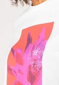 Camiseta de algodón blanca con un gran gráfico de flor rosa vibrante sobre un fondo coral. Cuello redondo, mangas cortas, textura suave.