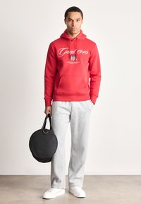 Homme portant un sweat à capuche rouge avec du texte blanc, un pantalon de survêtement gris, des baskets blanches, tenant un sac noir rond dans une main, se tenant contre un mur uni.