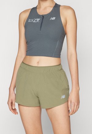 Femme portant un crop top de sport gris sans manches avec une demi-fermeture éclair et un short de sport vert olive, debout devant un fond uni.