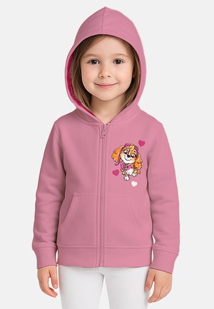 Young child wearing a pink zip-up hoodie with a cartoon dog and hearts graphic on the chest, paired with white pants.