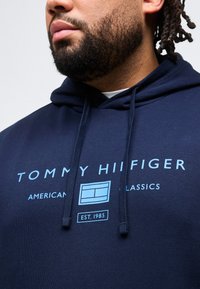 Άνδρας με γενειάδα που φοράει σκούρα μπλε φούτερ με κουκούλα, με ανοιχτό μπλε κείμενο "Tommy Hilfiger American Classics Est. 1985" και λογότυπο στο στήθος.