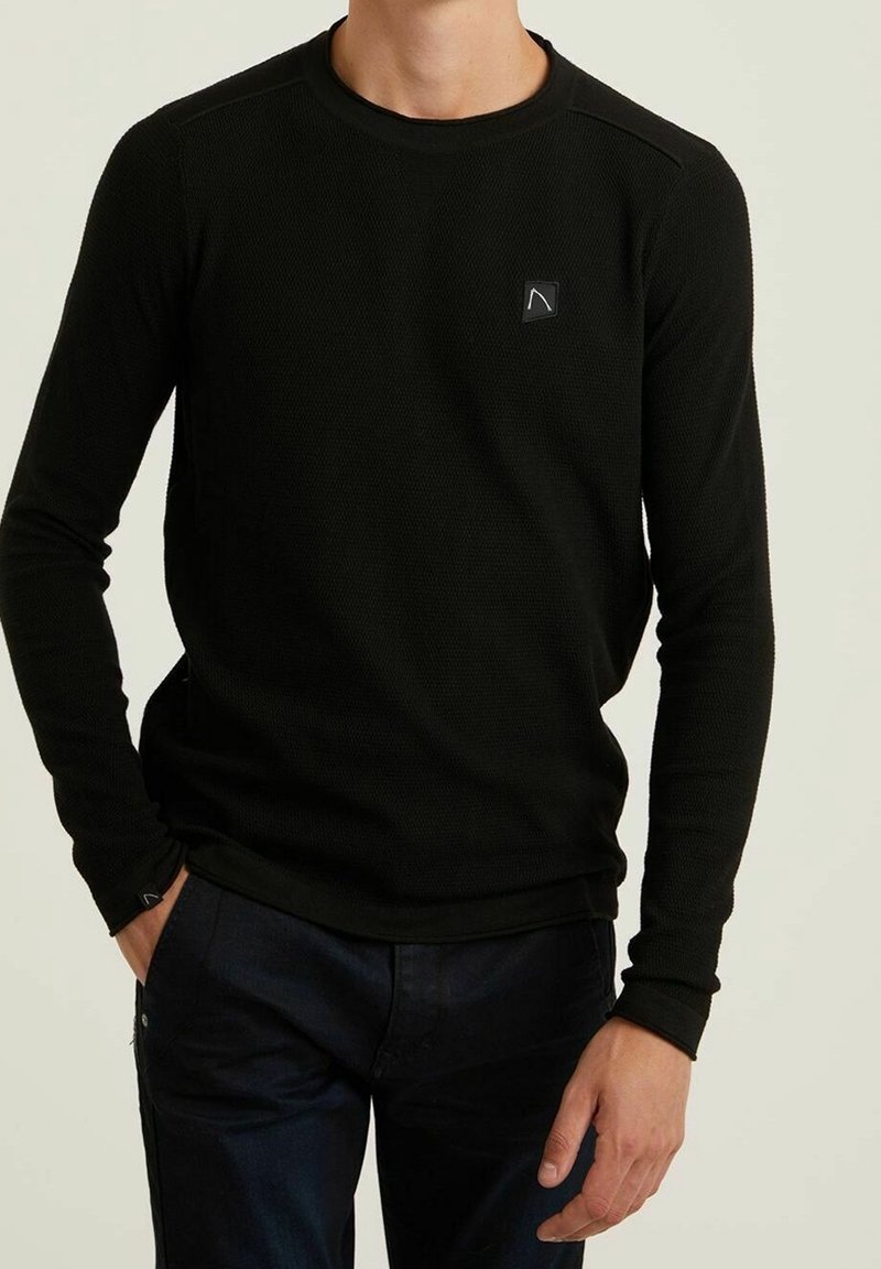 Homme portant un pull noir ajusté à manches longues avec un petit logo sur la poitrine gauche et un pantalon foncé, main dans la poche.