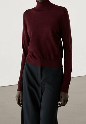 Personne portant un pull col roulé bordeaux ajusté et un pantalon noir taille haute à jambes larges sur un fond clair uni.