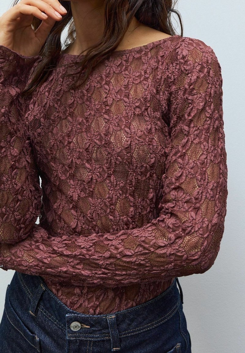 Femme portant un haut en dentelle mauve transparent à manches longues avec des motifs floraux, assorti à un jean taille haute bleu foncé, sur un fond uni.