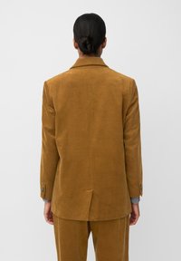 Bruine corduroy blazer met een gestructureerd ontwerp, enkele rugventilatie en knoopmanchetten, gezien van achteren tegen een effen achtergrond.