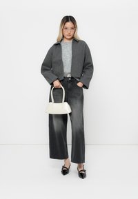 Giovane donna con occhiali indossa una giacca grigia, un maglione grigio, jeans a gamba larga neri e scarpe nere, mentre tiene in mano una borsa bianca su uno sfondo bianco.