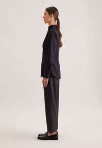 Camicia nera con bottoni e colletto alto, abbinata a pantaloni scuri su misura e mocassini neri. Tessuto liscio, taglio strutturato, design minimalista.