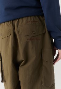 Pantaloni cargo verde oliva con due tasche posteriori e chiusure a bottone. Presentano testo ricamato rosso. Indossati con una maglietta a maniche lunghe blu navy.