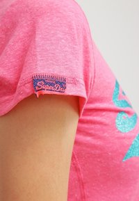 Camiseta de manga corta rosa con acabado texturizado y un logo de purpurina azul. Etiqueta cosida en la manga en colores contrastantes.