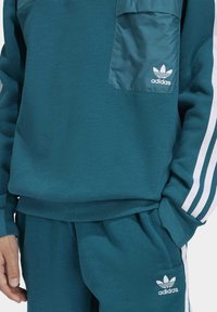 Chándal teal de Adidas con franjas blancas en las mangas y los pantalones, con logotipos de Adidas en el bolsillo del pecho y en la parte superior del muslo.