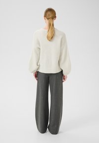 Lagani, kremasti pleteni pulover s balonskim rukavima i zaobljenim rubom, u kombinaciji s širokim tamno sivim hlačama koje imaju suptilne vertikalne pruge.