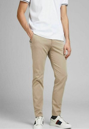 Homme portant un pantalon beige slim, un polo blanc avec des bordures noires, et des baskets blanches avec des rayures noires et rouges, debout avec une main dans la poche.