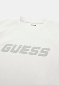 Balta džemperis su apvali iškirpte ir raglano rankovėmis. Pilkai atspausdintas "GUESS" logotipas iškarto matomas krūtinės srityje. Minkšta audinio tekstūra.