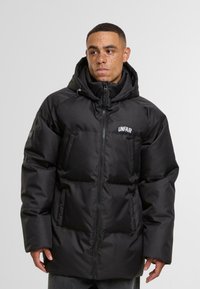 Svart boblejakke med glidelås foran, stor hette og "UNFAIR" logo. Har polstrede seksjoner, sidelommer og en glatt, blank tekstur.