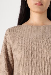 Pull en tricot beige avec une large encolure bateau, présentant une texture côtelée et des manches amples, mettant en avant un aspect doux et confortable.