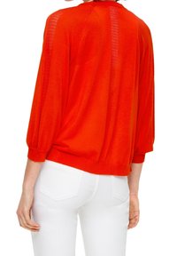 Czerwony sweter dzianinowy z rękawami o długości do połowy ramienia i bufkami, z teksturowanym panelem z tyłu. Noszony z białymi spodniami, prezentując luźny fason.