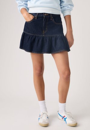 Persoană care poartă o fustă mini din denim închis cu tiv cu volane, bluză cu mâneci lungi de culoare albastru deschis, șosete albe și adidași albi cu dungi albastre și roșii.