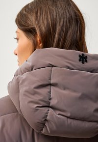 Chaqueta de plumas marrón con un collar alto, que presenta un logo negro brillante en la parte posterior. El material parece suave con secciones cosidas.