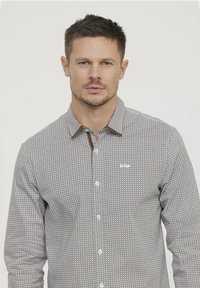 Chemise grise à motifs avec de petits designs géométriques, boutons blancs et accent marron au col. Présente un logo sur la zone gauche de la poitrine.