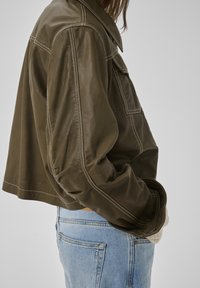 Persoană purtând o jachetă de piele maro cu cusături albe și blugi deschis la culoare, stând cu mâinile în buzunarele jachetei, pe un fundal simplu.