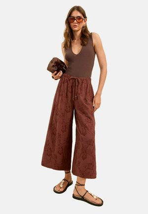 Naine, kes kannab pruuni varrukateta toppi, laiade säärega lillemustrilisi pükse, suureid päikeseprille, kandes kootud käekotti ning kannab kestadega kaunistatud sandaale.